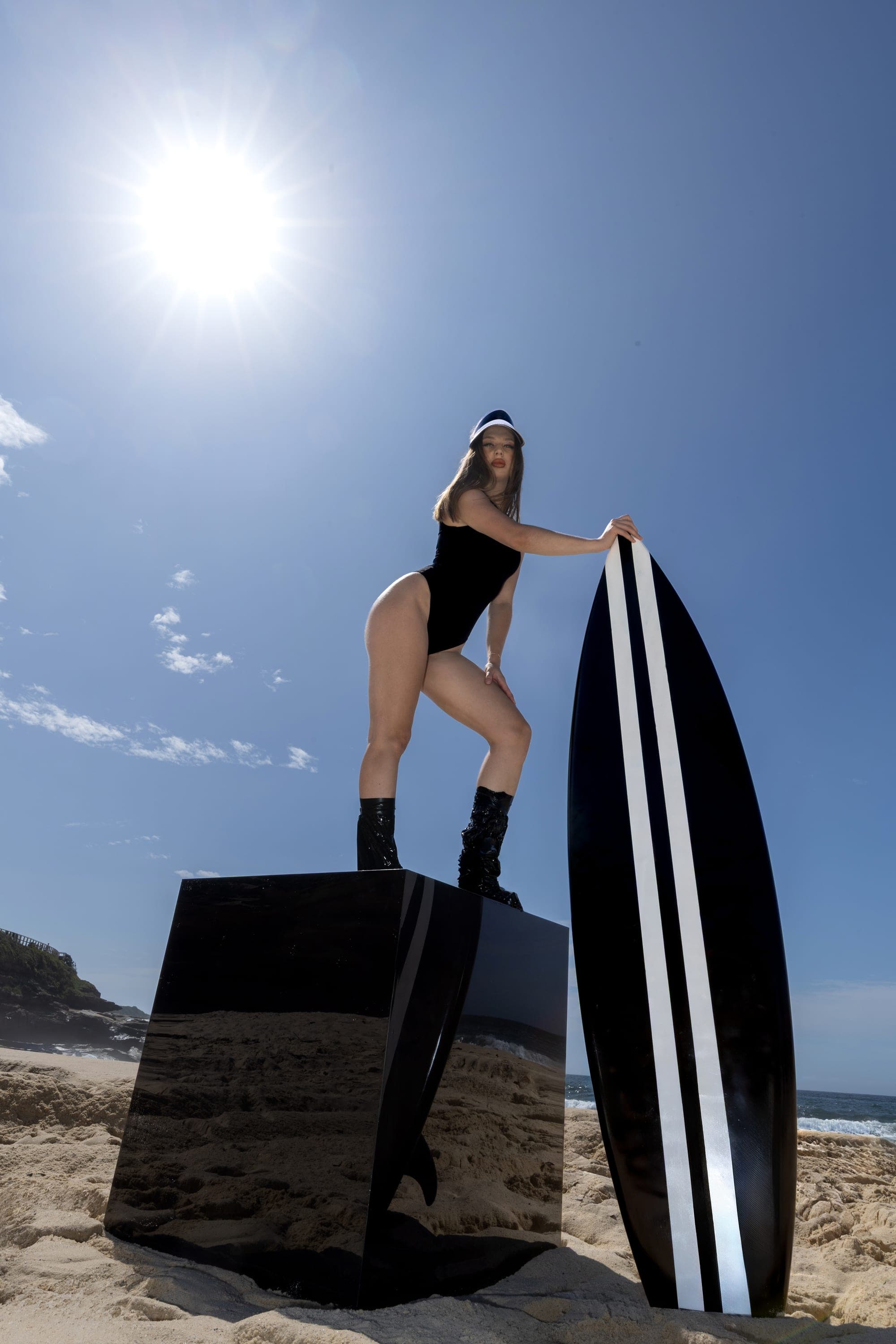 Model in a black swimsuit and visor leaning on a surfboard, Sydney beach fashion photography
