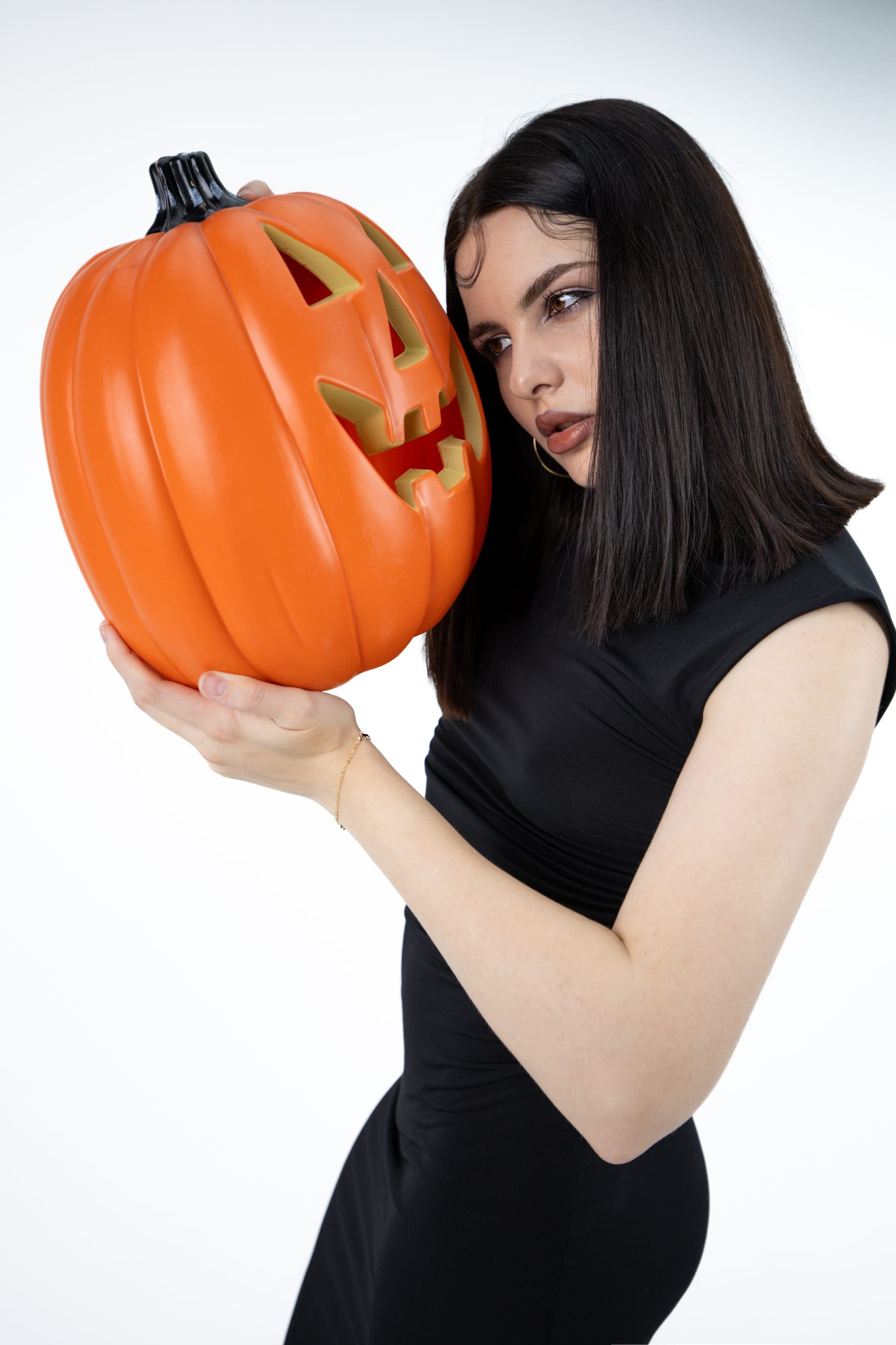Model in a black dress with a jack-o-lantern prop on a white studio backdrop, Halloween-themed shoot