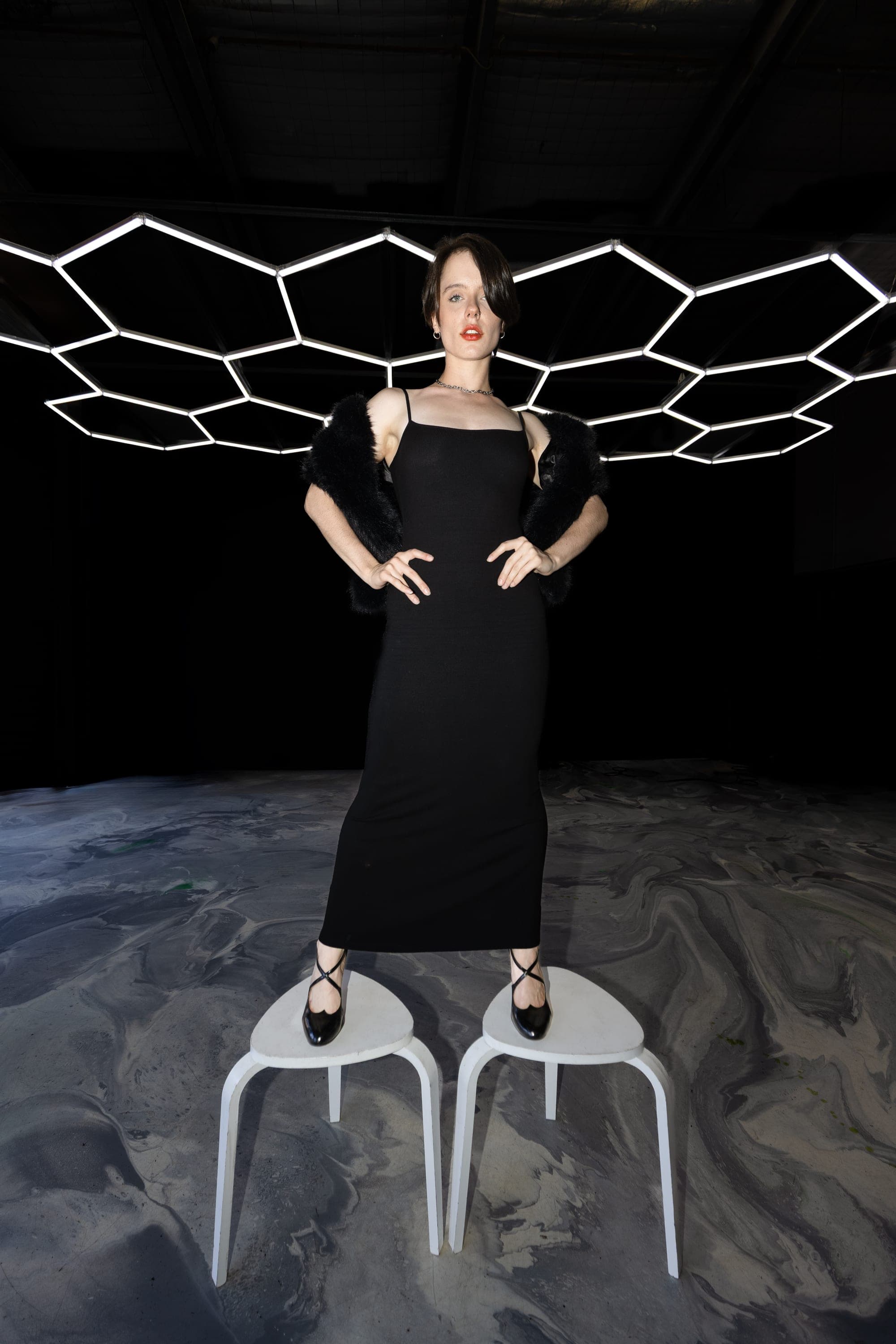 Model in a black slip dress and faux fur stole under a geometric light installation, studio photography